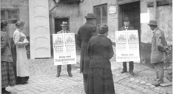 Svartvitt foto av två personer som håller i valaffischer och en grupp människor som tittar på.