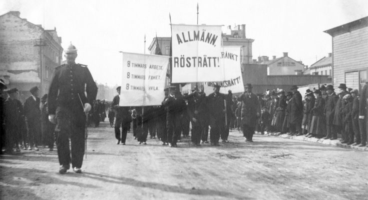 Svartvitt fotografi av män som går med fanor där det står allmän rösträtt och 8 timmar arbete.