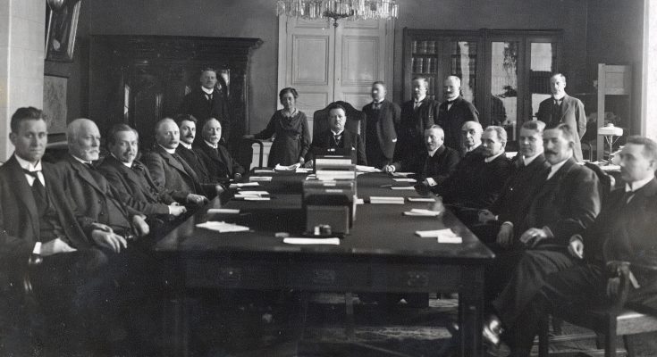 Svartvitt foto av ett femtontal män kring ett bord och en kvinna som står upp bredvid sin stol.