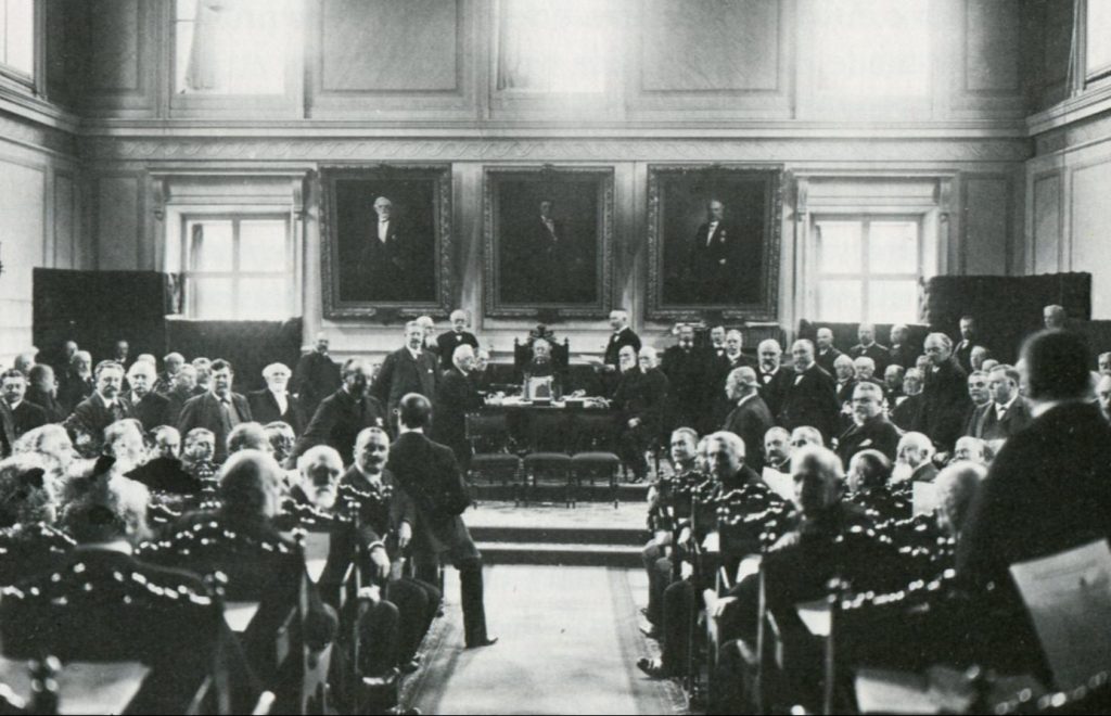 Ett svartvitt fotografi av många män i en sal som vänder sig om och tittar mot kameran.