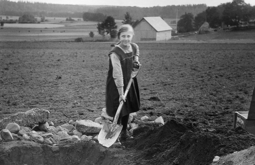 Ett svartvitt fotografi av en flicka som håller i en spade vid en åker.