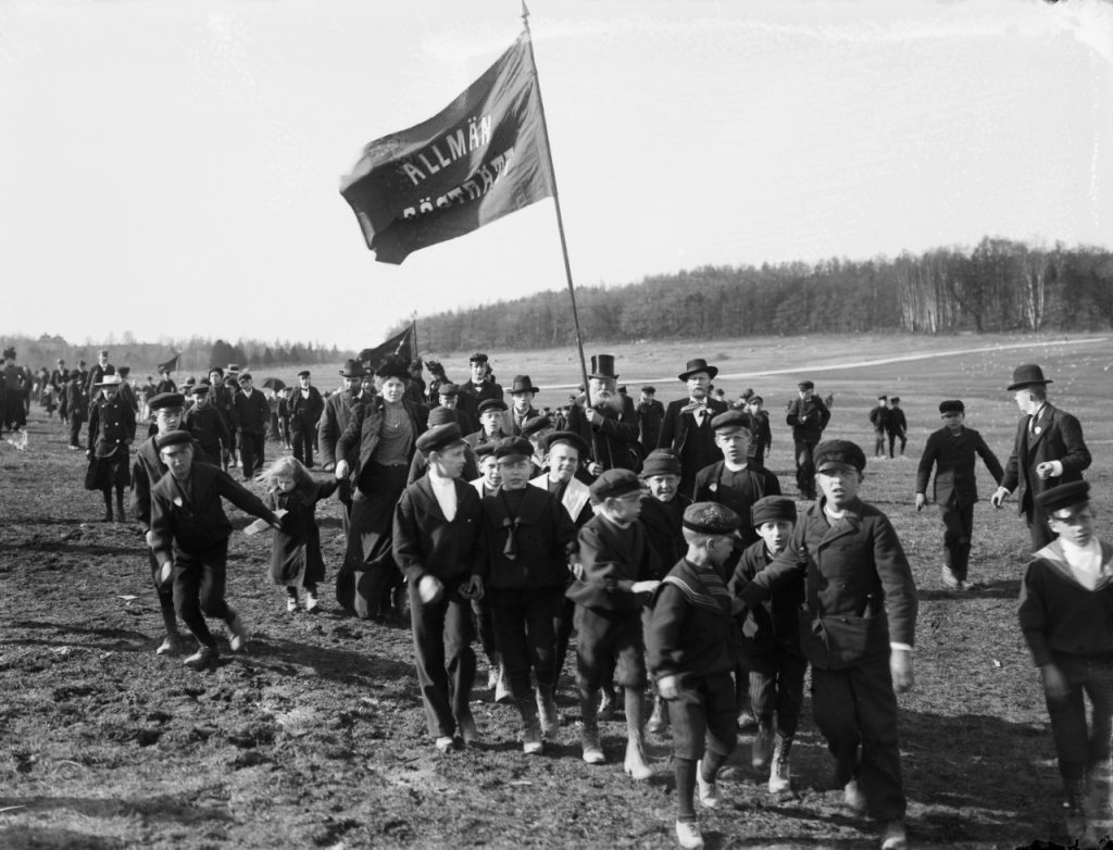 Svartvitt foto av ett demonstrationståg med en fana med texten Allmän rösträtt.