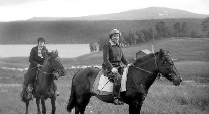 Svartvitt foto av två kvinnor på hästar i ett landskap med fält och berg.