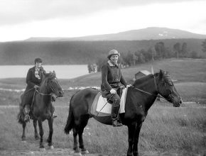 Svartvitt foto av två kvinnor på hästar i ett landskap med fält och berg.