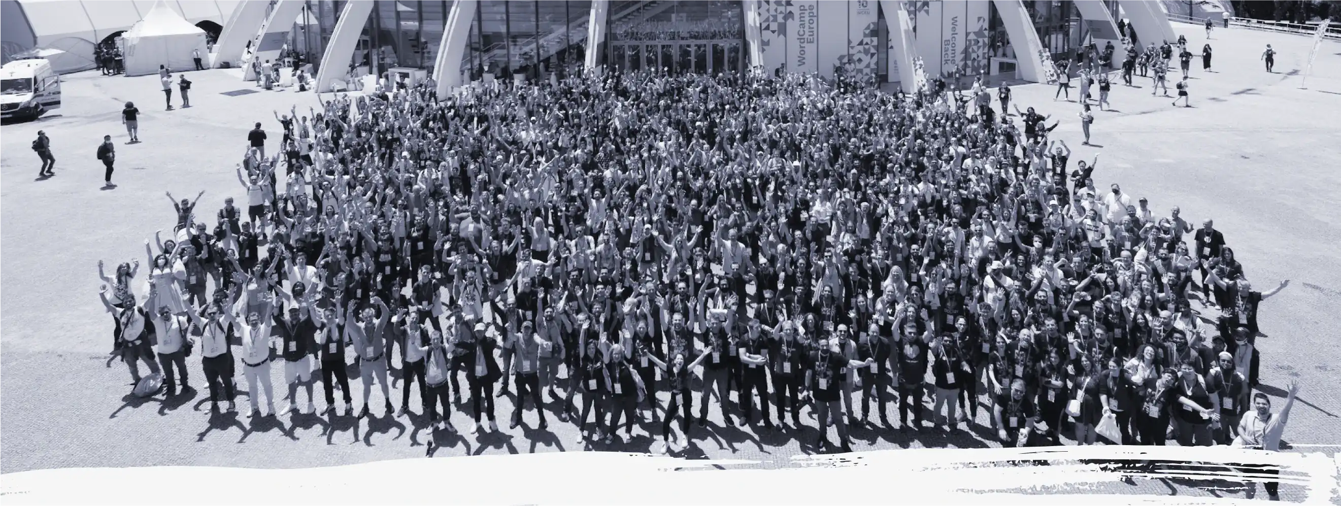 Una vista d'ocell en blanc i negre dels milers de col·laboradors que van assistir a la WordCamp Europe 2022. Tots estan drets davant del recinte amb les mans aixecades en senyal de celebració.